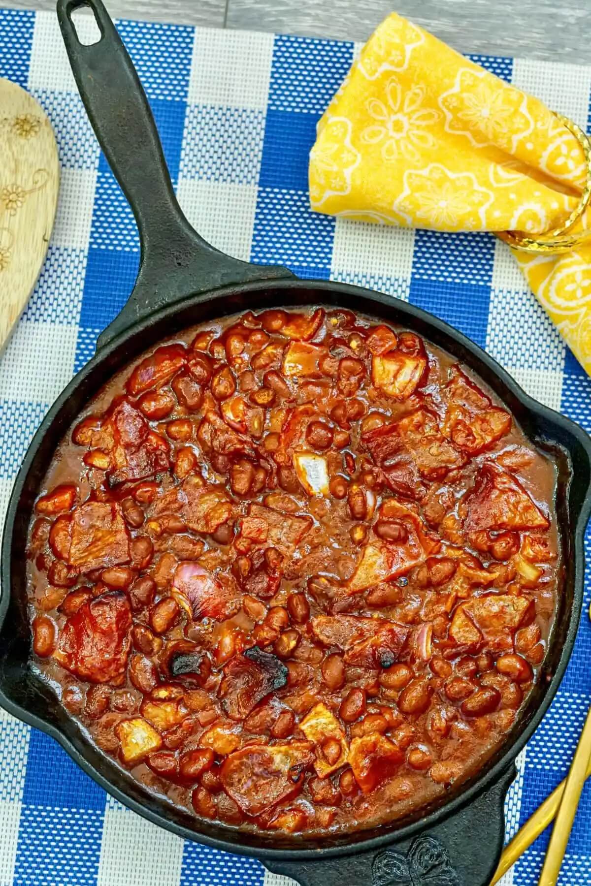 iron skillet on a blue check tablecloth with baked beans and bacon