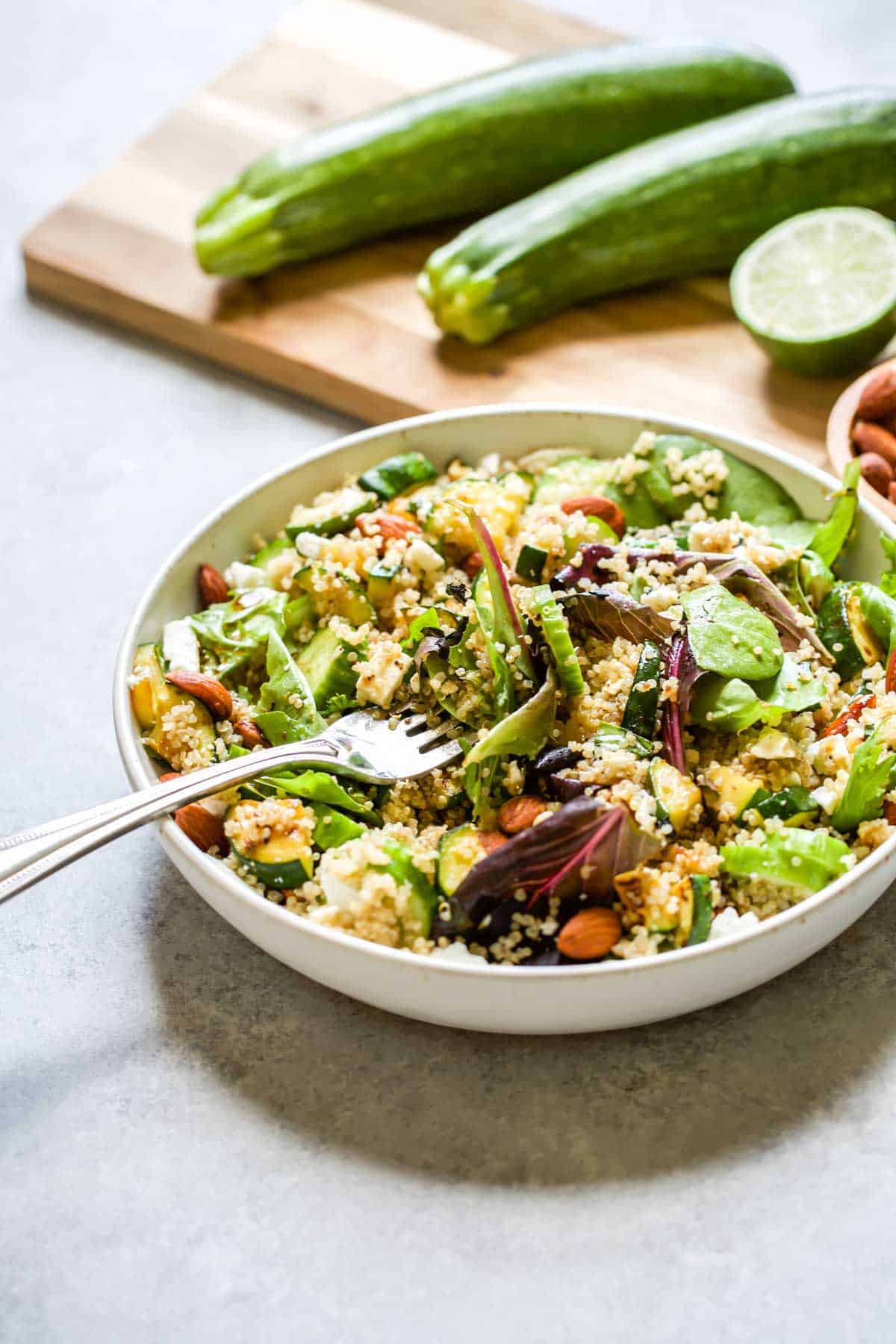 bowl of quinoa salad with fork, wooden chopping board with cucumbers in background