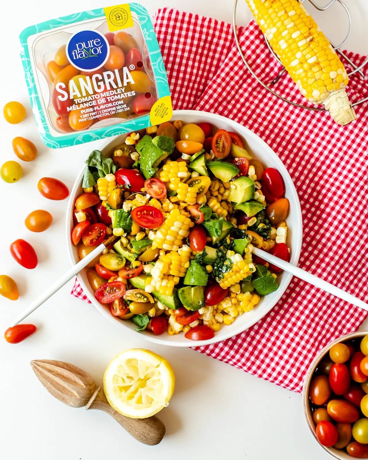 bright coloured corn salad in a white bowl with salad tongs, sitting on a red checked cloth and extra tomatoes to the side