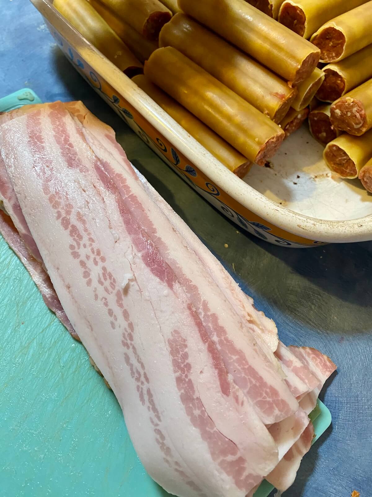slices of bacon on a chopping board next to a tray of stuffed cannelloni shells.