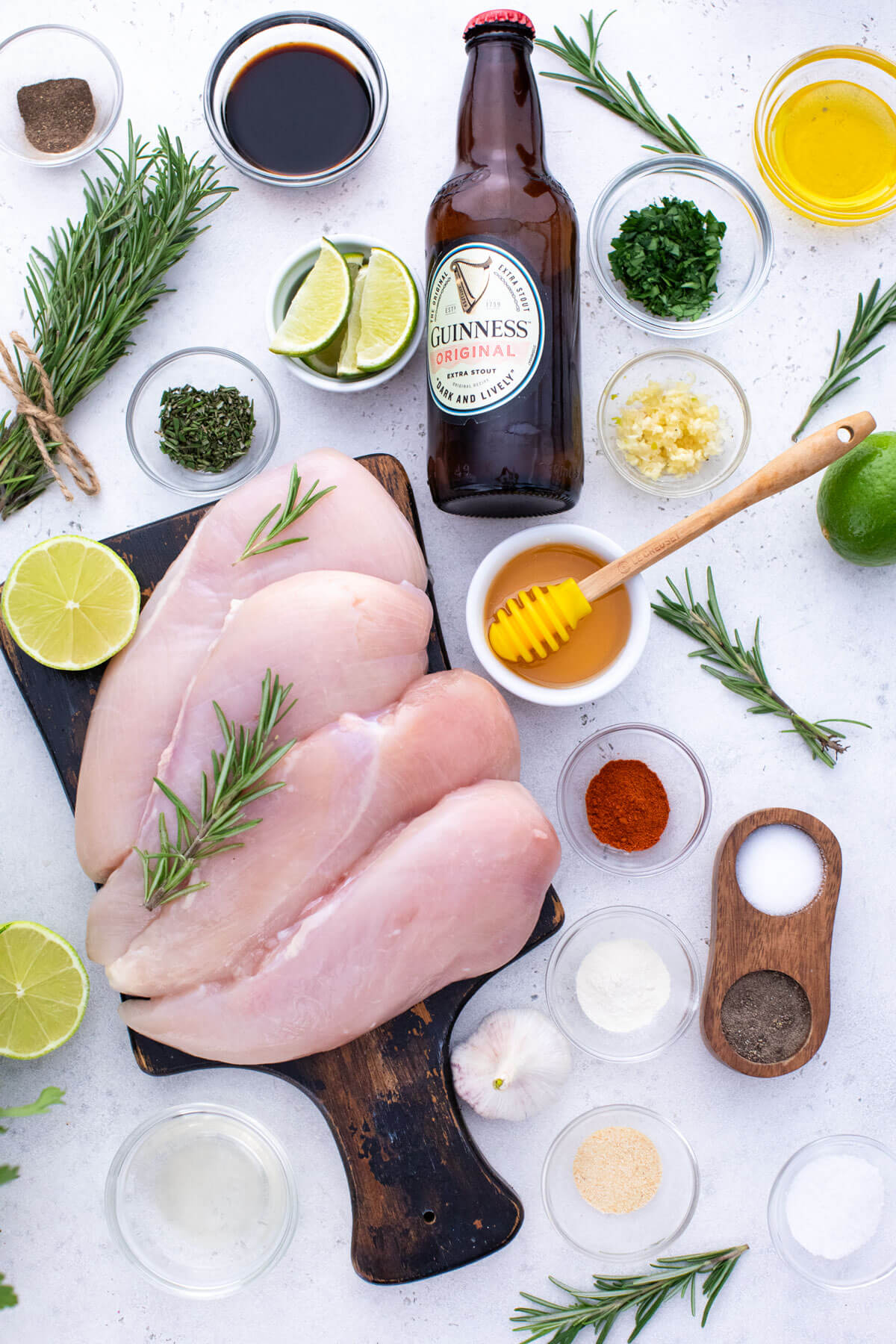 ingredients for grilled guinness chicken in a flat lay format with chicken breasts on wooden serving board, bottle of guinness beer and other ingredients in small bowls.