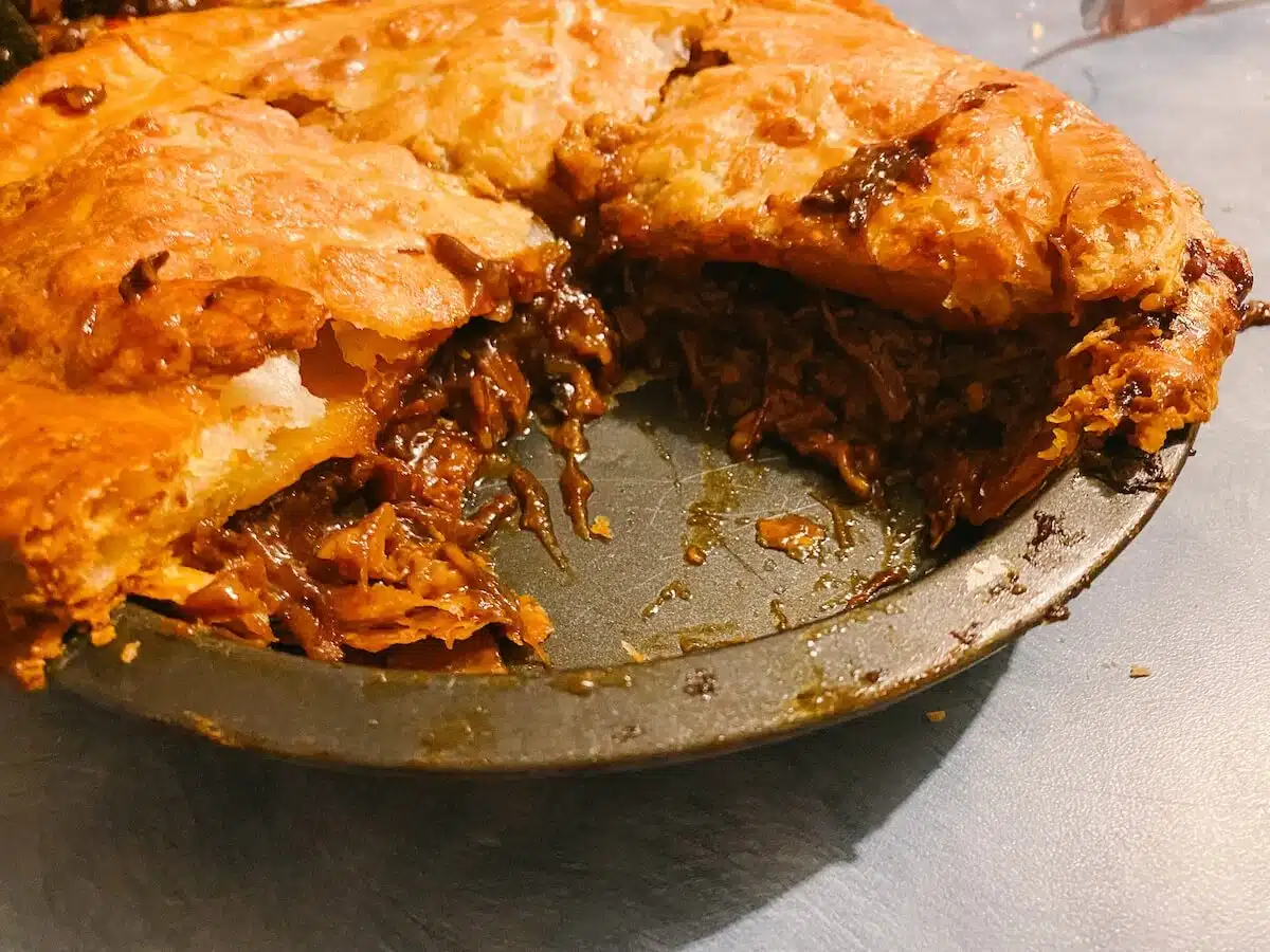 A close-up of a savory pie with a golden-brown crust. A large slice has been removed, revealing a filling of tender, shredded meat and rich gravy. The pie sits on a dark metal pie dish, and the filling is slightly spilling over the edge onto the dish.