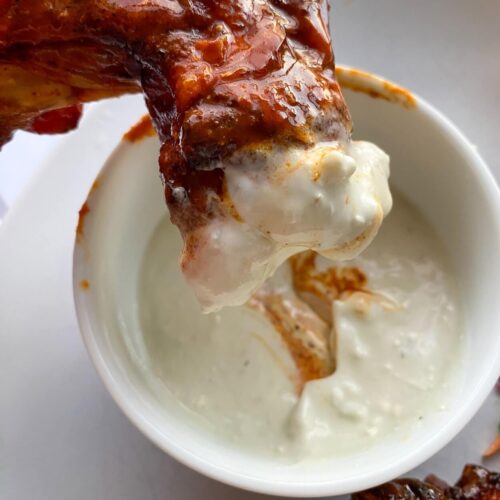 dipping a chicken wing into a small white bowl of creamy blue cheese sauce.