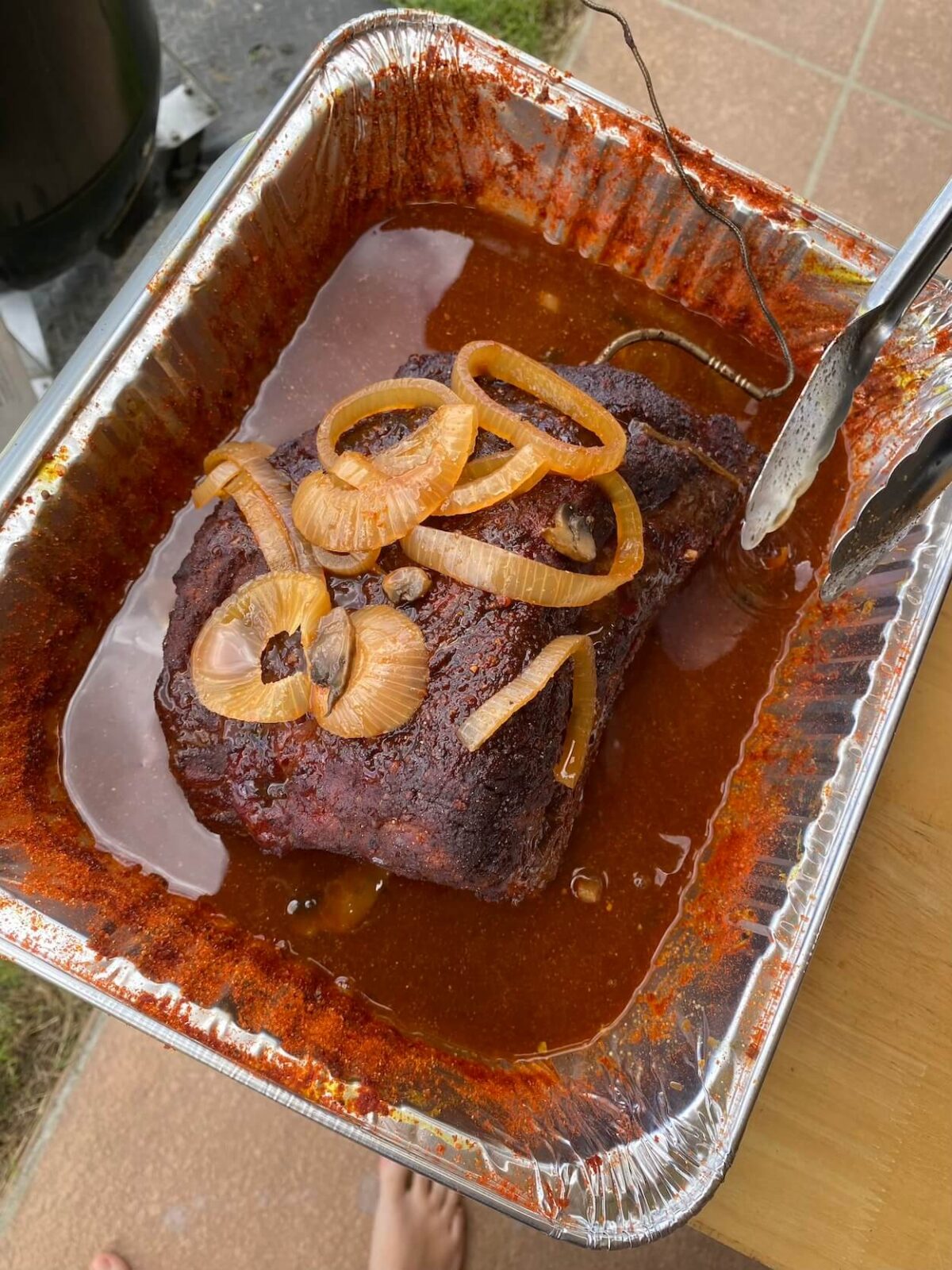 semi-smoked brisket sitting in tray of braising liquid with cut onions on top.